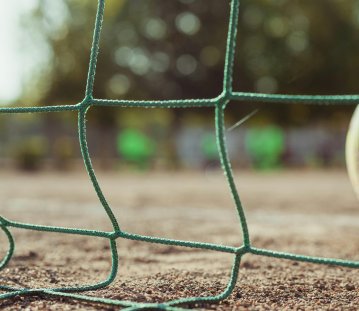 Nahaufnahme eines grünen Fußballnetzes, das im Vordergrund scharf zu sehen ist. Im unscharfen Hintergrund erkennt man einen Ball am rechten Bildrand und eine Spielfläche im Freien.