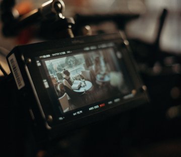 Eine professionelle Filmkamera zeigt auf ihrem Monitor eine Filmszene, in der eine Frau an einem Tisch sitzt und in einem Buch liest. Die warme, stimmungsvolle Beleuchtung und der unscharfe Hintergrund vermitteln einen cineastischen Blick hinter die Kulissen einer Filmproduktion.