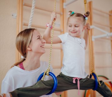 Ein Kind mit weißem T-Shirt und dunkler langer Hose trainiert an Gymnastikringen, während eine Lehrerin es aufmerksam begleitet und motivierend anlächelt.