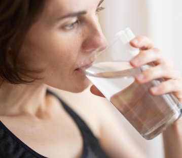 Eine Frau mit schulterlangen, dunklen Haaren und einem schwarzen Tanktop trinkt ein Glas Wasser