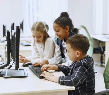 Zwei Mädchen und ein Junge im Grundschulalter sitzen in einem Computerraum gemeinsam vor einem Bildschirm, der Junge tippt etwas auf der Tastatur
