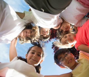 Eine Gruppe von Kindern steht im Kreis und blickt lächelnd nach unten in die Kamera. Die Aufnahme ist von unten gemacht, sodass der Himmel und etwas Baumlaub im Hintergrund zu sehen sind. Die Kinder wirken fröhlich und halten vermutlich zusammen oder lehnen sich zueinander.