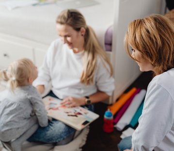 Eine Frau, wie eine weitere Frau mit einem kleinen Kind gemeinsam ein Bilderbuch anschaut. Die drei sitzen in einer hellen, wohnlichen Umgebung und wirken aufmerksam miteinander im Gespräch.