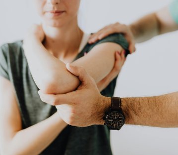 Eine Frau im dunkelgrünen T-Shirt (Gesicht ist nicht zu sehen) wird von einem Physiotherapeuten an der Schulter behandelt, indem er eine Hand auf ihrer Schulter hat und die andere an ihrem Ellenbogen.