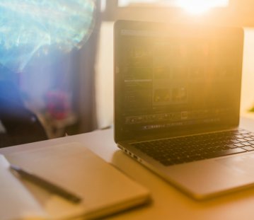 auf einem hellen Schreibtisch liegen ein Notizbuch mit einem Stift links und recht ein aufgeklappter Laptop, auf dem verschwommen ein dunkler Bildschirm mit Zahlen zu erkennen ist. Im Hintergrund scheint durch das Fenster die Sonne und eine Spiegelung der Sonne verdeckt den restlichen Hintergrund.
