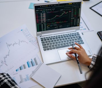 Person analysiert Börsencharts auf einem Laptop und vergleicht diese mit ausgedruckten Diagrammen. Die Szene zeigt die Auswertung von Finanz- und Marktdaten.
