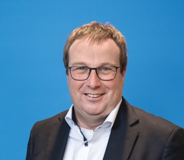 Ein Mann mit braunen kurzen Haaren, Brille, weißem Hemd und schwarzem Jackett lächelt in die Kamera