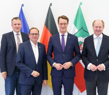 Von links nach rechts stehen Bürgermeister Tobias Stockhoff, Oberbürgermeister Thomas Kufen, Ministerpräsident Hendrik Wüst und Landrat Dr. Olaf Gericke gemeinsam vor den Flaggen der Europäischen Union, Deutschlands und Nordrhein-Westfalens. Die vier Politiker tragen dunkle Anzüge und blicken in die Kamera.