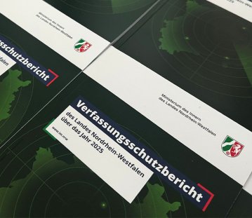Mehrere Exemplare eines gedruckten Berichts liegen nebeneinander auf einem Tisch. Auf den Titelseiten steht „Verfassungsschutzbericht des Landes Nordrhein-Westfalen über das Jahr 2025“ sowie „Ministerium des Innern des Landes Nordrhein-Westfalen“ mit Landeswappen. Das Design zeigt eine grüne Karte mit kreisförmigen Markierungen auf dunklem Hintergrund.