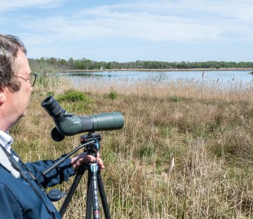 Minister Krischer steht am Rand eines Schilfgebiets und beobachtet mit einem Spektiv auf einem Stativ eine Wasserfläche. Im Hintergrund sind ein See, Vögel und eine flache, bewaldete Landschaft unter blauem Himmel zu sehen.