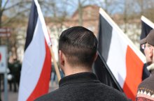 verschiedene Personen stehen mit dem Rücken zum Bild und Fahnen in der Hand zusammen. Die Fahne hat einen schwarzen, einen weißen und einen roten Streifen und symbolisiert die Reichsflagge