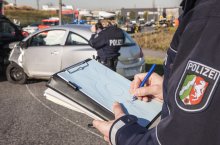 Ein Polizist hält ein Klemmbrett in der Hand, auf dem er einen Unfallhergang aufzeichnet. Im Hintergrund fotografiert eine Polizistin zwei Autos, die zusammengestoßen sind, dahinter ist ein Stau zusehen.