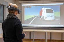 eine VR-Brille tragende Frau steuert einen digitalen LKW vor einer Leinwand