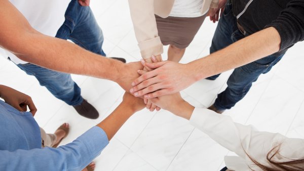 5 Menschen stehen im Kreis und haben jeweils ihre linke Hand in der Mitte sternförmig übereinander gelegt als Symbol für gute Teamarbeit.
