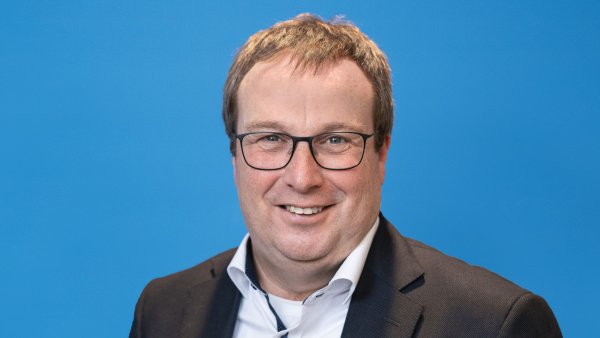 Ein Mann mit braunen kurzen Haaren, Brille, weißem Hemd und schwarzem Jackett lächelt in die Kamera