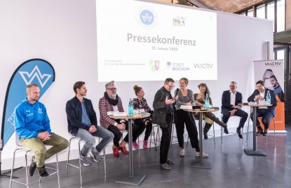 Staatssekretärin Andrea Milz sitzt mit Teilnehmern der Pressekonferenz auf Hockern an Rundtischen. Hinter ihnen eine weiße Rückwand mit einer Präsentation.