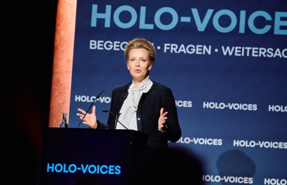 Eine Frau in dunklem Blazer steht gestikulierend an einem Redner-Pult 