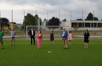 Staatssekretärin MIlz mit Vereinsmitgliedern vor dem Tor eines Rasen-Fußballplatzes. Sie in rosapinker Sportbekleidung mit einem Fußball in der Hand.