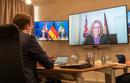 Ein Mann am Schreibtisch schaut auf zwei Bildschirme an der Wand. Im linken sind zwei Männer, im rechten eine Frau vor Flaggen zu sehen.