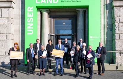 Eine Gruppe Menschen steht vor einem Eingang mit grünem Tor, zwei halten jeweils eine große Karte