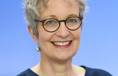 Eine Frau mit kurzem grauen Haar und Brille vor blauem Hintergrund