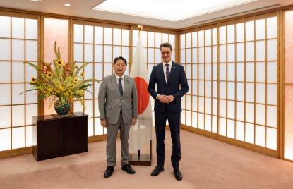 Zwei Männer in Anzügen stehen in einem japanischen Raum mit Japan-Flagge und einer Kommode und Blumen im Hintergrund