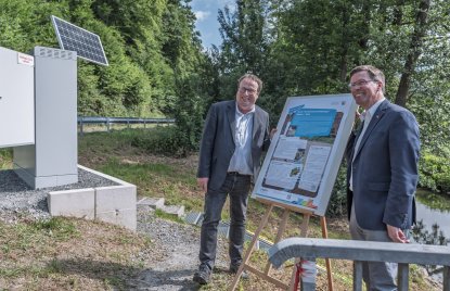 Minister Krischer und ein weiterer Mann im Anzug stehen neben einer Tafel, auf der verschiedene Stichpunkte zu den Hochwassermeldepegeln aufgeführt sind. Die Tafel steht am neuen Hochwassermeldepegel in Welzen, von dem im Hintergrund eine Treppe zu sehen ist.
