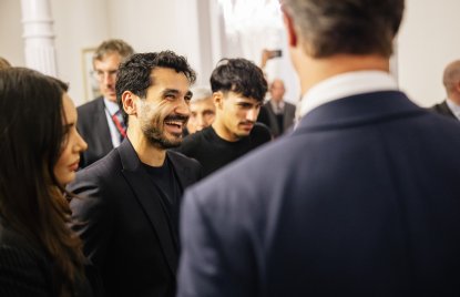 Fußballer İlkay Gündoğan trägt einen dunklen Anzug und lacht zwischen anderen Personen in Business-Kleidung.