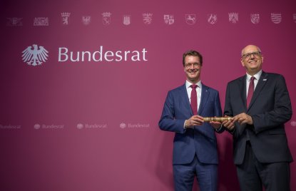 Zwei Männer in Anzügen halten gemeinsam einen Holzstab fest.
