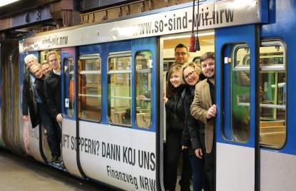 Ein Gruppe Menschen schaut aus einer U-Bahn, die mit der Werbung der Finanzverwaltung NRW verziert ist