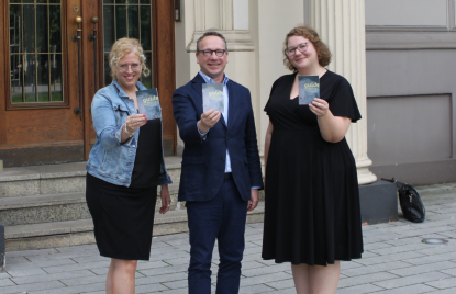 Zwei Frauen und ein Mann stehen vor einer Eingangstür und halten ein Ratgeberheft hoch