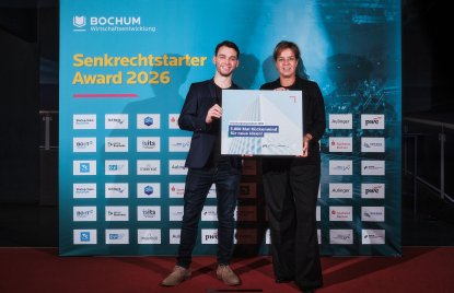 Zwei Personen präsentieren gemeinsam den „Senkrechtstarter Award 2026“ vor einer Sponsorenwand mit Logos und der Aufschrift „BOCHUM Wirtschaftsentwicklung“. Rechts ist Ministerin Mona Neubaur zu sehen.