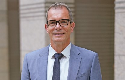 Ingo Dudenhausen steht im blauen Anzug und dunkelblauer Krawatt und weißem Hemd vor beigen Säulen. Er trägt eine Brille und lächelt frontal in die Kamera.