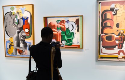 Eine Person bepackt mit Taschen und gezückter Kamera steht vor drei bunten Gemälden an der Wand