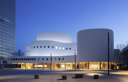 Das Düsseldorfer Schauspielhaus bei Dämmerung mit beleuchteter, geschwungener weißer Fassade und markanten Baukörpern. Im Vordergrund liegt ein weitläufiger Platz, flankiert von Hochhäusern.