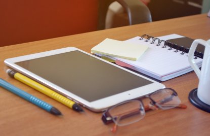 Auf einer Tischplatte liegen zwei Stifte, eine Brille, ein Tablet und ein Notizbuch
