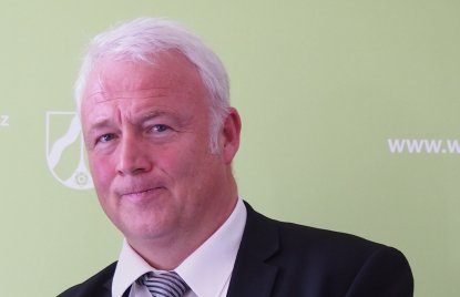 ein Mann mit grauem Haar, dunklem Sakko und gestreifter Krawatte steht vor einer grünen Wand