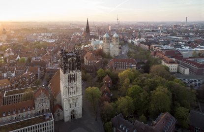 Blick von oben auf Münsters Altstadt: Im Vordergrund steht die Überwasserkirche, dahinter ist der Dom mit seinen beiden Türmen und die Lambertikirche zu sehen. Am Horizont erkennt man Münsters höchstes Bauwerk, den Fernsehturm. 