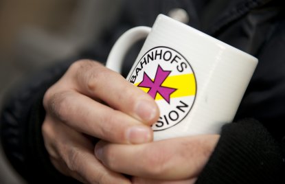 zwei Hände halten einen weißen Becher mit dem Logo der Bahnhofsmission darauf