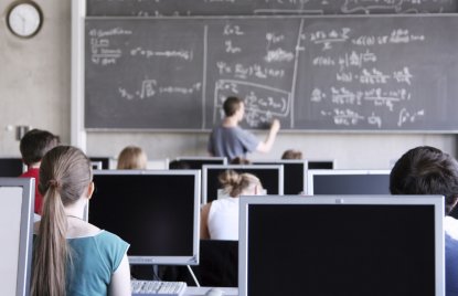 Auf dem Bild zu erkennen sind Schülerinnen und Schüler in einer Unterrichtsstunden am PC. Ein Lehrer schreibt Formeln an die Tafel.