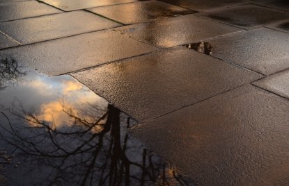 auf einem Bürgersteig spiegeln sich ein Baum und Wolken in einer Pfütze