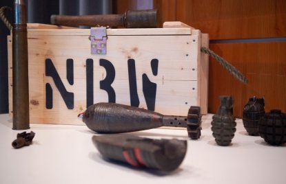 Holzkiste mit der Aufschrift „NBW“ und verschiedenen historischen Sprengkörpern und Granaten auf einem Ausstellungstisch. Die Objekte sind vor einem warm beleuchteten Hintergrund arrangiert und wirken wie Exponate in einer Ausstellung oder Sammlung.