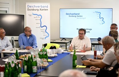 Umweltminister Oliver Krischer sitzt in einem weißen Hemd an einem Konferenztisch und spricht mit mehreren Personen. Im Hintergrund sind ein Bildschirm und ein Banner des Deichverbands Duisburg-Xanten zu sehen.