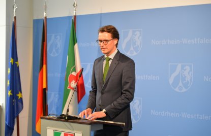 Verkehrsminister Wüst vor blauer NRW-Wand an einem Rednerpult, links neben ihm die Flaggen von Europa, Deutschland und NRW.