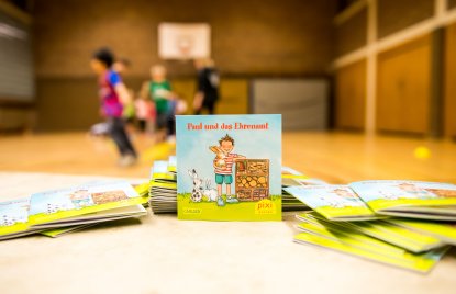 Ein Pixi Buch steht aufgestellt auf einem Tisch. Im Hintergrund sieht man Kinder,die in einer Sporthalle spielen.
