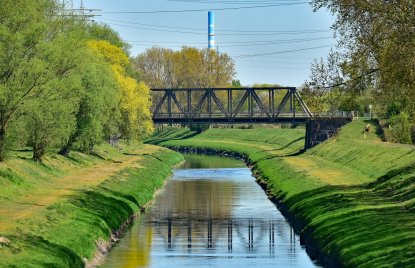 Blick auf einen begrünten Kanal in Oberhausen mit einer Stahlbrücke, spiegelndem Wasser und Bäumen entlang der Ufer. Im Hintergrund sind Stromleitungen und ein markanter Industrieschornstein zu sehen