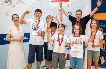 5 Kinder und 3 Erwachsene jubeln, die Kinder haben Medaillen an roten Bändern um den Hals