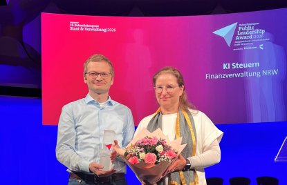 Zwei Personen stehen auf einer Bühne und präsentieren einen Preis sowie einen Blumenstrauß vor einer großen Leinwand. Im Hintergrund ist die Auszeichnung für das Projekt ‚KI Steuern‘ der Finanzverwaltung NRW beim Zukunftskongress Staat & Verwaltung 2026 zu sehen.