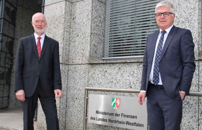 Zwei grauhaarige Männer in Anzügen stehen vor dem Schild des Finanzministeriums