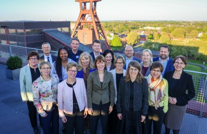 Gruppenfoto von etwa 15 Personen in formeller Kleidung auf einer Dachterrasse, die in die Kamera lächeln. Im Hintergrund steht ein großes Fördergerüst aus Stahl sowie Gebäude und eine grüne Landschaft. Die Personen stehen in zwei Reihen dicht beieinander.